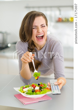 Stock Photo: Laughing young woman eating salad and watching tv in kitchen