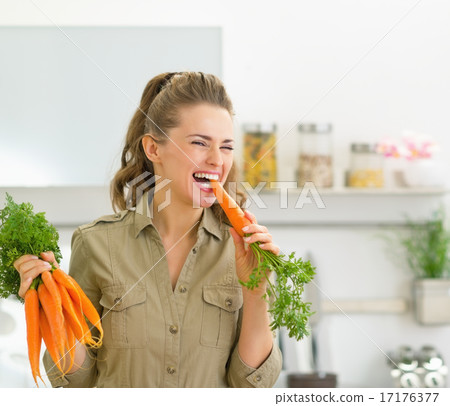 Happy young housewife eating carrot in kitchen 17176377