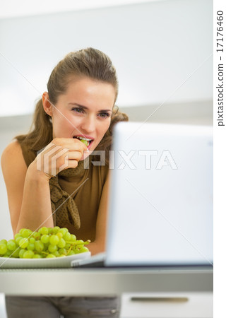 Happy young housewife eating grape and looking in laptop Happy young housewife eating grape and looking in laptop 17176460