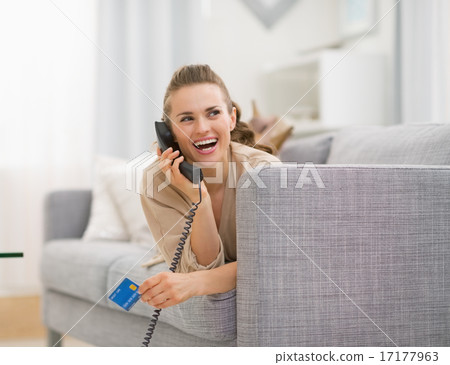 Happy young woman with credit card laying on sofa and talking ph 17177963