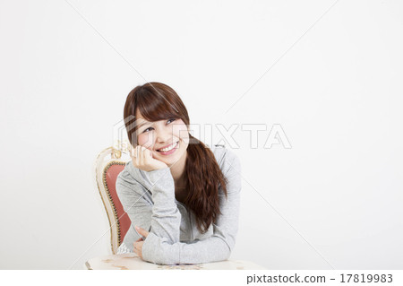 與坐在白色背景前面的一把椅子和cheeking關於她的右手的一張微笑的面孔的年輕微笑在桌上 17819983