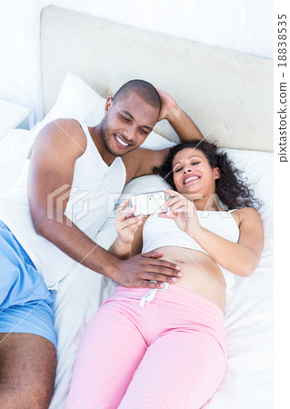 Pregnant wife holding smartphone with husband lying on bed 18838535