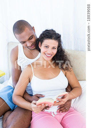 Portrait of pregnant wife holding book with husband on bed 18839088