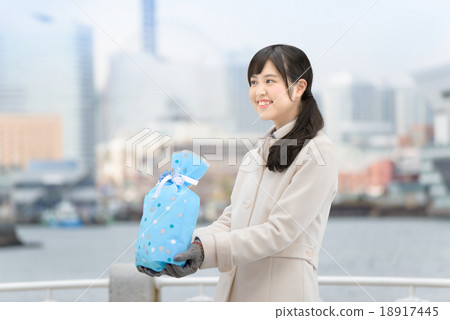 A young woman standing wearing a coat with a gift in a park where the sea can be seen 18917445