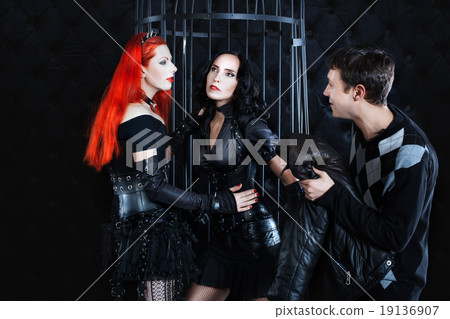 Two women dressed in fetish play, the man looks. Two women dressed in fetish play, the man looks. 19136907