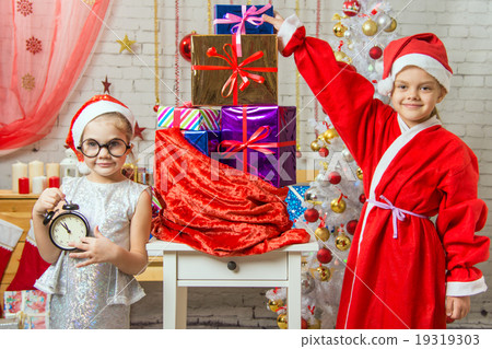 A girl holding a clock, a girl dressed as Santa Claus is standing at the bag with gifts 19319303