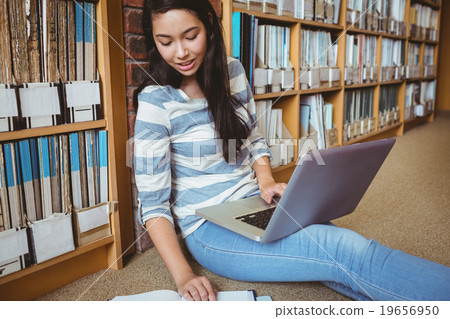 Smiling student sitting on the floor against wall in library studying with laptop and books 19656950
