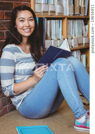Smiling student sitting on the floor against wall in library reading book 19656951