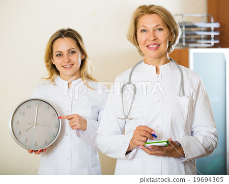 Two friendly doctors holding clock and smiling in modern clinic 19694305