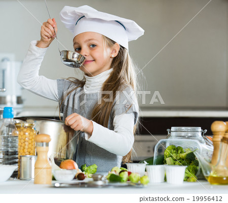 Smiling female child in chef cap making bouillon in kitchen Smiling female child in chef cap making bouillon in kitchen 19956412
