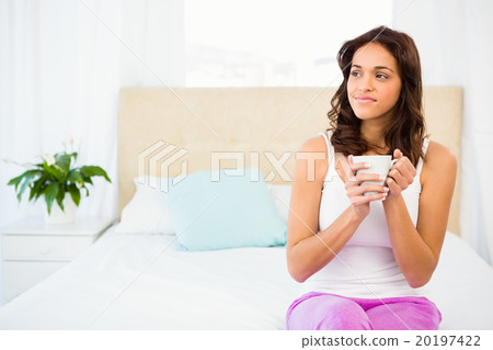 Happy woman holding cup of coffee while sitting on bed 20197422