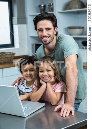 Smiling father using laptop with his children in kitchen Smiling father using laptop with his children in kitchen 20198646