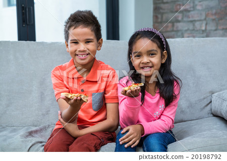 Smiling siblings eating pizza on the sofa 20198792