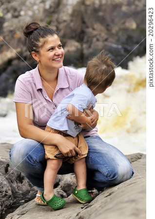 Cheerful young woman holding her little son near raging river on background 20296423