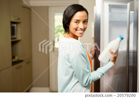 Smiling brunette holding milk bottle with open refrigerator 20579200