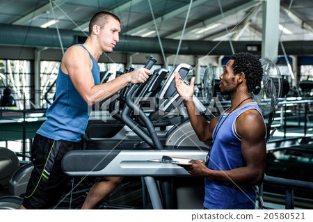 Muscular man using elliptical machine with trainer Muscular man using elliptical machine with trainer 20580521