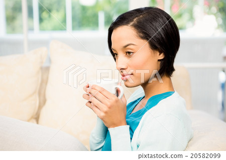 Attractive brunette holding cup while sitting on the sofa Attractive brunette holding cup while sitting on the sofa 20582099