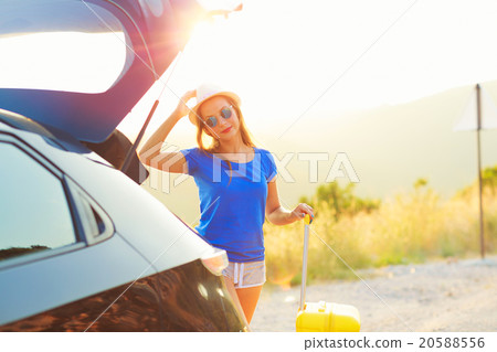 Woman with a yellow suitcase standing near the trunk of a car pa 20588556