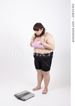 A fat young woman holding a towel on her neck in front of a white background and holding it with both hands and watching the scale 20655302