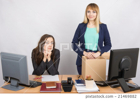 The girl in the office is facing the box with things near the colleague sitting next 21469882