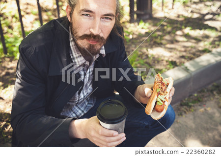Asian man with hot dog and coffee in park 22360282