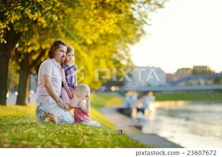 Young father and his daughters sitting by a river Young father and his daughters sitting by a river 23607672