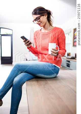 Young woman with mobile phone and mug sitting on table Young woman with mobile phone and mug sitting on table 23971116