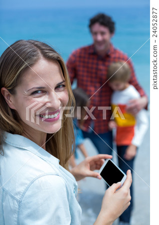 Cheerful mother holding cellphone while family in background Cheerful mother holding cellphone while family in background 24052877