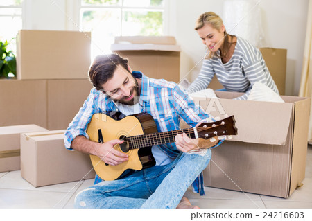 Man playing a guitar while woman unpackaging cardboard boxes in background Man playing a guitar while woman unpackaging cardboard boxes in background 24216603