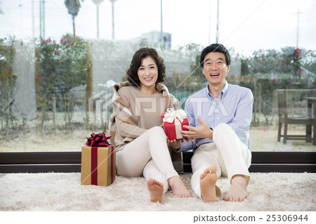 Middleaged Asian Couple Smiling and Sitting with Gift Boxes on Rug In Livingroom Middleaged Asian Couple Smiling and Sitting with Gift Boxes on Rug In Livingroom 25306944