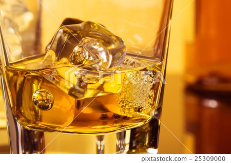 detail of glass of whiskey with ices near bottle on table with reflection, warm atmosphere detail of glass of whiskey with ices near bottle on table with reflection, warm atmosphere 25309000