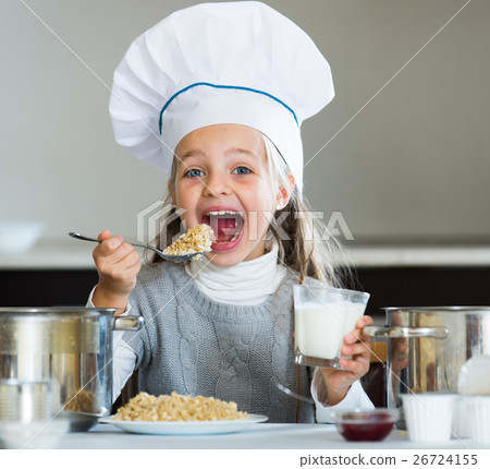 Cheerful girl in cook cap eating porridge indoors Cheerful girl in cook cap eating porridge indoors 26724155