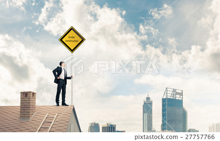 Young businessman on house brick roof holding yellow signboard a 27757766