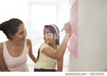 Smiling mother looking at her daughter writing message on refrigerator Smiling mother looking at her daughter writing message on refrigerator 28231881