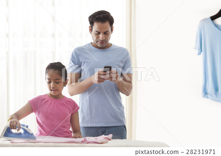 Daughter ironing clothes and father using mobile phone Daughter ironing clothes and father using mobile phone 28231971
