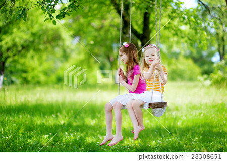 Two cute little sisters having fun on a swing together in beautiful summer garden Two cute little sisters having fun on a swing together in beautiful summer garden 28308651