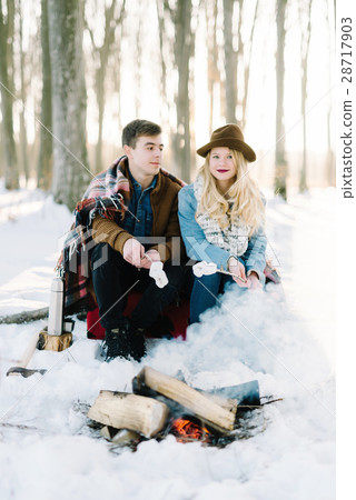stylishly dressed young couple, sitting near the fire in the winter forest stylishly dressed young couple, sitting near the fire in the winter forest 28717903