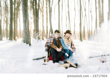 stylishly dressed young couple, sitting near the fire in the winter forest stylishly dressed young couple, sitting near the fire in the winter forest 28717904