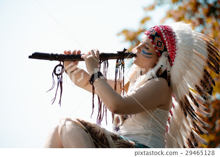 Young woman in war bonnet headdress of American Indian plays on a tree a flute 29465128