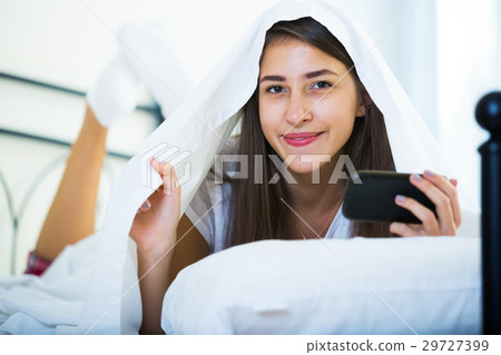 Happy girl sending sms under blanket indoors Happy girl sending sms under blanket indoors 29727399
