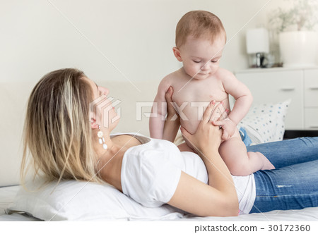Baby boy in diaper sitting on mother lying on bed Baby boy in diaper sitting on mother lying on bed 30172360