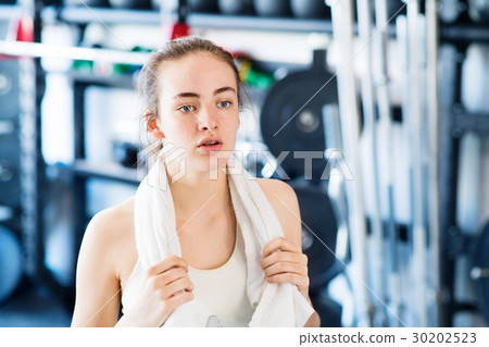 Young woman in gym, towel around her neck, resting Young woman in gym, towel around her neck, resting 30202523