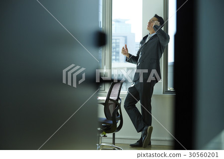The businessman leaning against the wall is talking on cell phone. 30560201