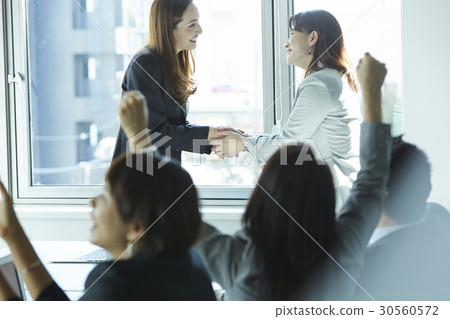 Two businesswomen are shaking hands and a group of people is cheering 30560572