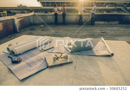 Construction equipment such as blueprint, Safety glasses and technology laptop over the photo blurred of engineers working with sun ray and lens flare background, industrial concept, worm color tone 30685252