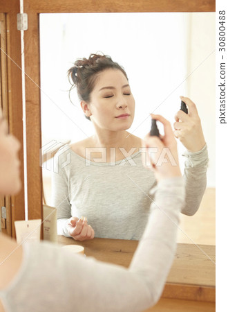 a woman is spraying lotion on her face at home. 30800488