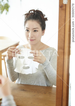 a woman sits in front of a mirror and lifts up facial mask sheet. 30800578