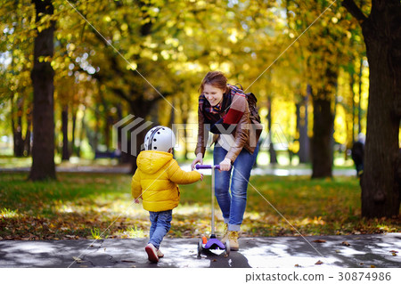 Young mother showing her toddler son how to ride a scooter in a autumn park Young mother showing her toddler son how to ride a scooter in a autumn park 30874986