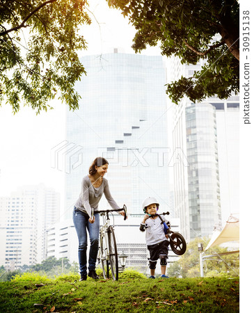 Little boy and his mother riding bicycle in the park with cityscape background 30915138