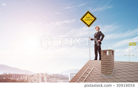 Young businessman on house brick roof holding yellow signboard and looking at city. Mixed media Young businessman on house brick roof holding yellow signboard and looking at city. Mixed media 30918058